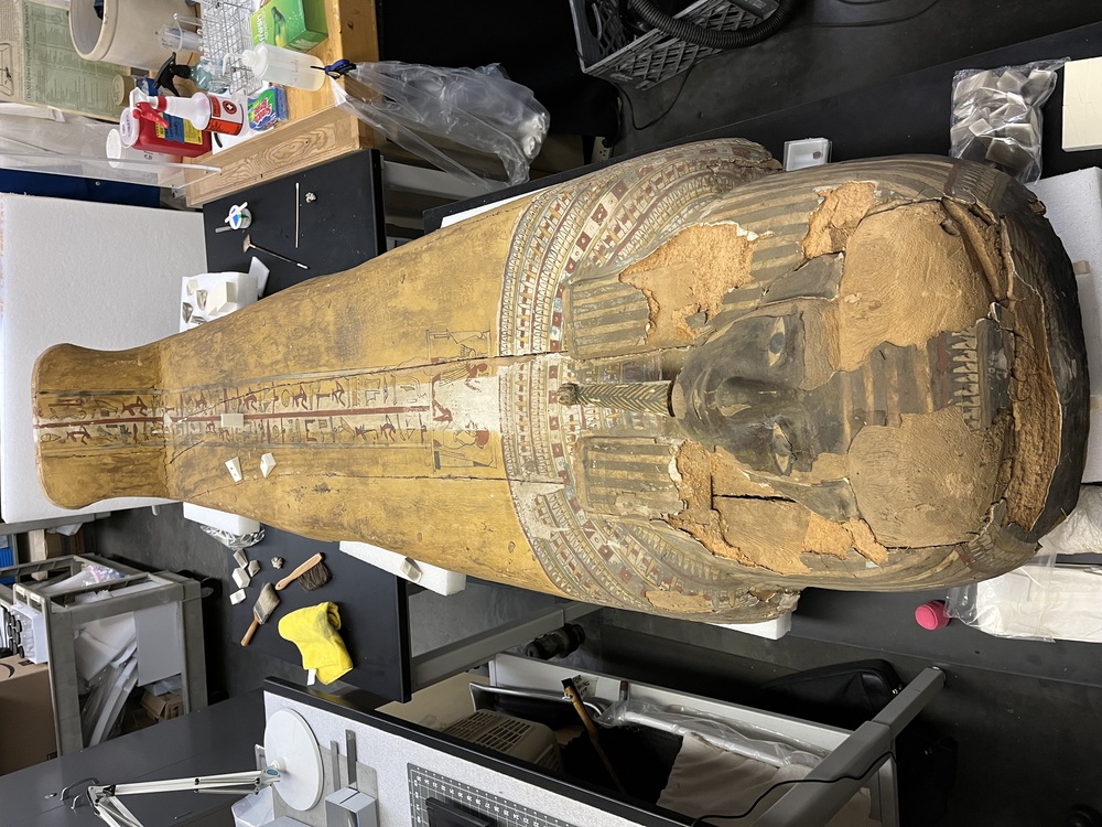 Hearst Museum object 122 of 167 titled Coffin, accession number 6-19912, described as Wooden anthropoid coffin. Inscription running down center of lid. Determined by Kea Johnston to probably be the outer coffin of Iwefaa (6-19928, 12-11039). According to Kea Johnston, "This coffin belonged to a man named Iwefaa, whose name is inserted in the middle of the spells written in the two columns on the front. This coffin is likely the outer coffin for PAHMA 6-19928 based on the similarity of the owner's name, unusual costume, and the paleography of the inscription. Both coffins were purchased on the antiquities market by George Reisner for Phoebe A Hearst. However, stylistically, they are very similar to a set of coffins known to have come from the site of Akhmim, which was excavated in the 1880s by Gaston Maspero, and which has heavily looted for the antiquities market during this decade." "The pectoral vignette between the text and the collar shows the deceased standing before squatting baboon and is the source of our logo. The hieroglyph for 'sky' is above their heads, and the side borders are formed by a pyramid and the symbol for the West.