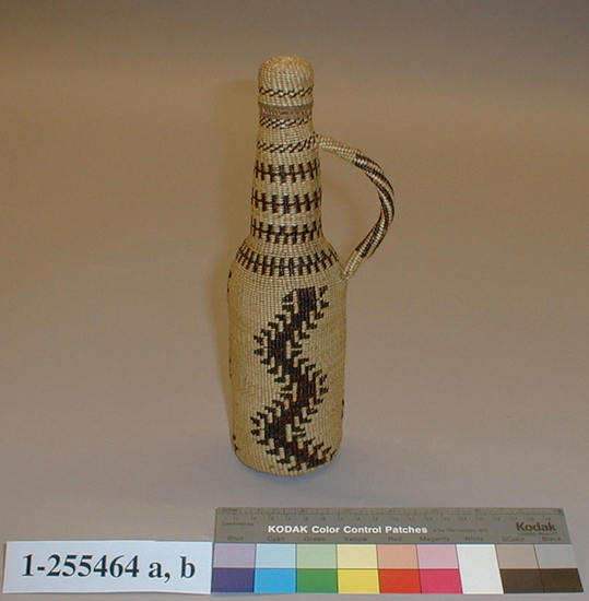 Hearst Museum object titled Bottle and lid, accession number 1-255464a,b, described as (a) Brown glass bottle with baskery covering. (b) Basketry lid and handle. Geometric design in dark brown on natural colored ground. Plain twined with full twist overlay, tule cordage warp, tule root and white grass overlay.