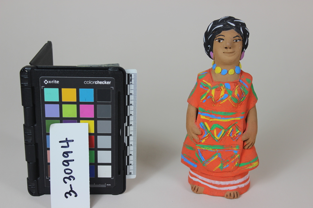 Hearst Museum object titled Figurine, accession number 3-30994, described as FEMALE FIGURE: Standing figure wearing three piece matching outfit, underskirt, short sleeved dress, apron. Orange with white, green, yellow, blue decoration; necklace of blue and yellow, purple earrings; Braided hair wrapped around head. Josefina Aguilar marked on inside and Made in Mexico on paper label inside.