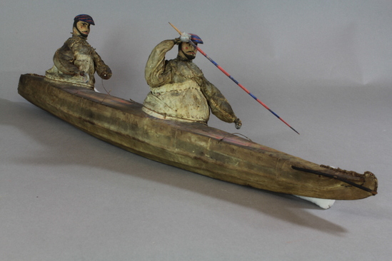 Hearst Museum object 6 of 8 titled Kayak model, accession number 2-4569, described as 2-hatch. Wood frame, rawhide cover. 2 human figures wearing hats painted bright blue and red and wearing sealskin parkas. 15 fixtures including weapons and paddles painted bright red and blue.