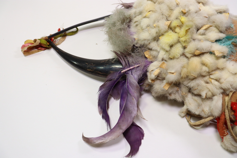 Hearst Museum object 2 of 15 titled Headdress, accession number 2-45410, described as Felt cap covered with strips of rabbit fur, patches stained yellow, traces of green fluff at front. Crown with 2 split polished horns attached to interior hide strip. Purple feathers and green-gray fluff in between and at base of horns. Multicolored silk grosgrain (?) ribbons bound to left horn tip. Ten "ermine tails" of rabbit fur sewn about a cord, red felt seized, with black hair tips; fringes at sides.