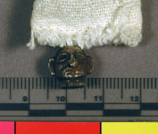 Hearst Museum object titled Bead, accession number 16-20458, described as metal (silver?) bead shaped like the heat of a man.  large ears, down turned mouth, large squinty eyes stringing hole from top of head through neck.