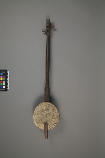 Hearst Museum object titled Musical instrument, accession number 18-1660, described as Musical instrument, stringed; scroll and pegs (for two steel strings) at top; gourd sounding box with sounding holes at bottom, covered with python skin; tail piece at bottom. Said to be old by collector. Length 66.5 cm. Diameter (gourd) 15.0 cm.