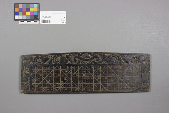 Hearst Museum object titled Board, accession number 18-1558, described as Calendar board, dark-stained wood; engraved board with multiple squares, some with circles within; diamond design down length of board; border on three sides has single eye and continuous floral design in deep relief. Augury board (”calenda”) for determining lucky and unlucky days for various enterprises. 30-day lunar months and seven day weeks. 37.7 x 11.4 cm.