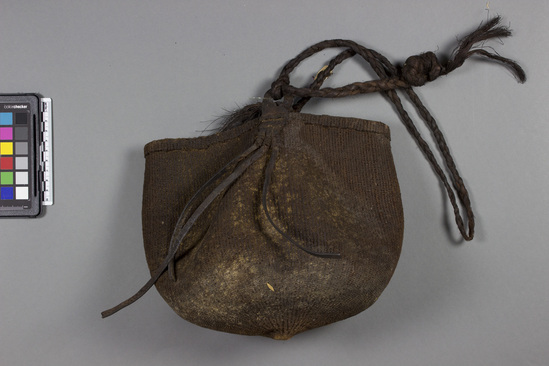 Hearst Museum object titled Bag, accession number 5-6945, described as String bag; twined black-brown vegetable fibre; braided twine strap attached to piece of leather sewn to both sides of bag; h. 17 cm; dia. 16 cm.