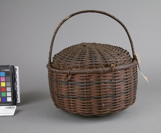 Hearst Museum object titled Basket, accession number 5-15645, described as Basket, fiber, plaited; round bottom; fixed arched carrying handle; dome shaped lid fitted to lip of bottom; hinged; dark brown; Rim diameter circa 24.5 centimeters X height (with lid) circa 18.5 centimeters.