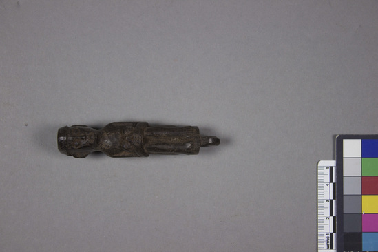 Hearst Museum object 1 of 2 titled Figurine, accession number 18-1480, described as Gurni figure, carved from dark brown wood; figure squats with hands clasped at knees; loop carved at bottom of feet; worn around neck. Worn around neck. Width 2.5 cm. Height 9.5 cm.