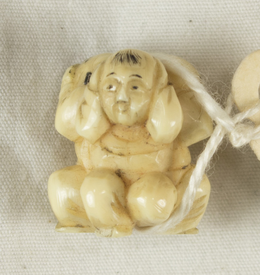 Hearst Museum object 10 of 10 titled Netsuke, accession number 9-12452, described as Japan. Ojime: carved ivory bead, of three boys crouched, backs together. One covers his ears, another eyes, and the last his mouth with hands ("Hear no evil, see no evil, speak no evil"). Surfaces angular. Some black paint detail. Height 2 centimeters. Hole runs through, between heads and feet.