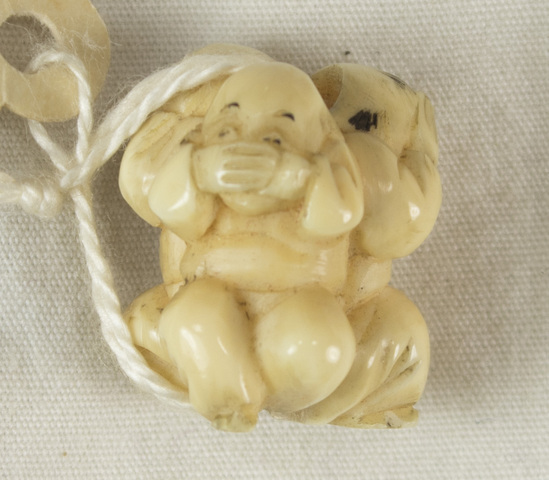 Hearst Museum object 7 of 10 titled Netsuke, accession number 9-12452, described as Japan. Ojime: carved ivory bead, of three boys crouched, backs together. One covers his ears, another eyes, and the last his mouth with hands ("Hear no evil, see no evil, speak no evil"). Surfaces angular. Some black paint detail. Height 2 centimeters. Hole runs through, between heads and feet.