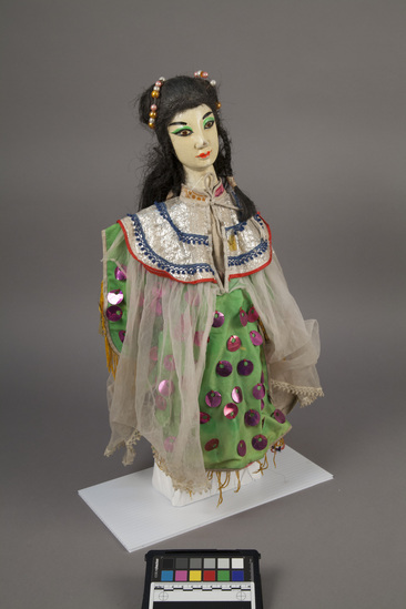 Hearst Museum object 3 of 6 titled Hand puppet, accession number 9-22235, described as Puppet, hand.  Foreign Princess A.  Head: carved wood, light orange with pink, green and black features, long, black hair with beads in it  B. Body: red and pink flowered print cloth,  Boots:  wood, black and white.  C. Dress: green cloth with purple sequins, sheer white overlay  48 x 31 cm (18 7/8 x 12 3/16 in)