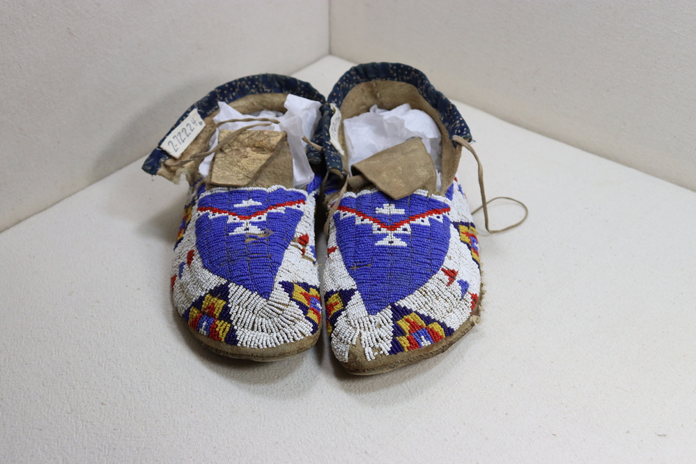 Hearst Museum object titled Beaded moccasins, accession number 2-72224a,b, described as a) beaded moccasins.  beaded thin leather upper stitched to heavier leather sole.  top of uppers trimmed in blue patterned cloth.  beaded patterns, mostly triangles, in blue, white, red, yellow, navy blue.  thin leather thong for tying. b) mirror image of a).