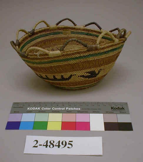 Hearst Museum object titled Basket, accession number 2-48495, described as Round with flat base and flaring sides. Raffia wrapped grass bundles form scalloped rim, circular design of canoes and ducks in dark blue with green stripes on staw-colored ground alternating with stripes of plaited cedar bark. Plain twined, white grass, natural and dyed, wrap twined, base with plaited and twined with cedar bark and tule. Attached tag states, "From Ye Olde Curiosity Shop, Coleman Dock Front, Seattle".