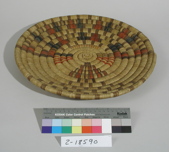 Hearst Museum object titled Plaque, accession number 2-18590, described as Basketry, coiled.  Kachina face in center of basket.  Elaborate headdress, brown, with red and blue filling; orange and green hatching on forehead.  Large brown eye with blue filler, opposite other brown eye with yellow filler.  Red hatchered triangle under each eye.  Mouth has vertical strips radiating out and down.  Checkerboard body.