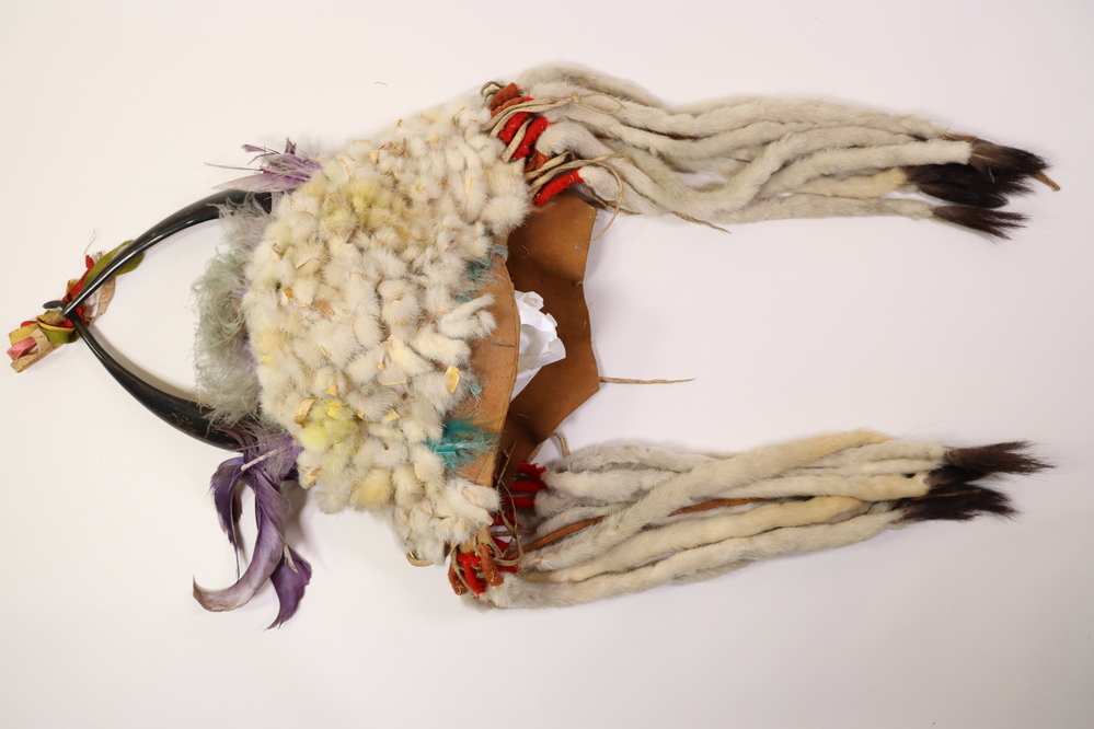 Hearst Museum object 13 of 15 titled Headdress, accession number 2-45410, described as Felt cap covered with strips of rabbit fur, patches stained yellow, traces of green fluff at front. Crown with 2 split polished horns attached to interior hide strip. Purple feathers and green-gray fluff in between and at base of horns. Multicolored silk grosgrain (?) ribbons bound to left horn tip. Ten "ermine tails" of rabbit fur sewn about a cord, red felt seized, with black hair tips; fringes at sides.