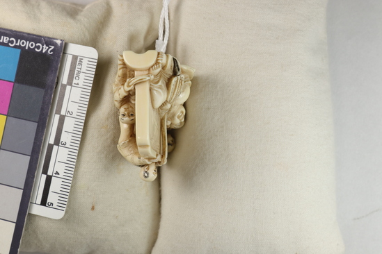 Hearst Museum object 13 of 14 titled Netsuke, accession number 9-18870, described as Netsuke; reclining figure of fat man facing to his right, supporting head on right hand; lying in front of wall; boy at left peering at man; boy at back of wall standing on barrel pulling at kimono cord; boy at right tugging at kimono hem; man holding fan in left hand; both sides of wall visible; H 3.6 cm X L 4.8 cm