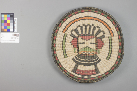 Hearst Museum object 3 of 3 titled Plaque, accession number 2-68500, described as Round, design showing kachina mask using black, red, green and orange on whitened, natural ground. Wicker weave, sumac warp, dyed rabbit brush weft, yucca for wrapped rim.