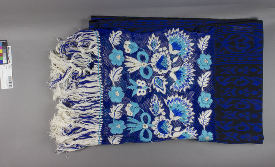 Hearst Museum object 2 of 2 titled Shawl, accession number 16-20211, described as Shawl; 76 x 145 cm, excluding knotting and fringe; ikat; black ground with blue ikat figures; starched royal blue knotting 35 cm l. with warp fringe plus added threads; embroidered flowres, leaves, and butterflies throughout knotted area in bright blue, turquoise, and white; silver sequins french knotted to flowers, leaves and butterflies;