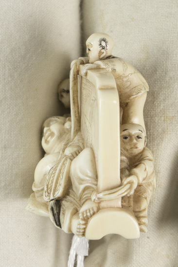 Hearst Museum object 14 of 14 titled Netsuke, accession number 9-18870, described as Netsuke; reclining figure of fat man facing to his right, supporting head on right hand; lying in front of wall; boy at left peering at man; boy at back of wall standing on barrel pulling at kimono cord; boy at right tugging at kimono hem; man holding fan in left hand; both sides of wall visible; H 3.6 cm X L 4.8 cm