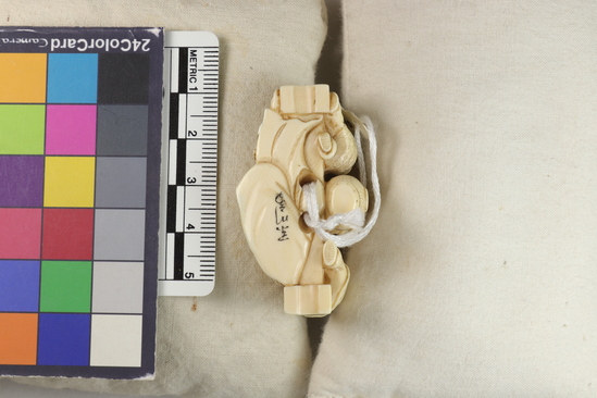 Hearst Museum object 5 of 14 titled Netsuke, accession number 9-18870, described as Netsuke; reclining figure of fat man facing to his right, supporting head on right hand; lying in front of wall; boy at left peering at man; boy at back of wall standing on barrel pulling at kimono cord; boy at right tugging at kimono hem; man holding fan in left hand; both sides of wall visible; H 3.6 cm X L 4.8 cm