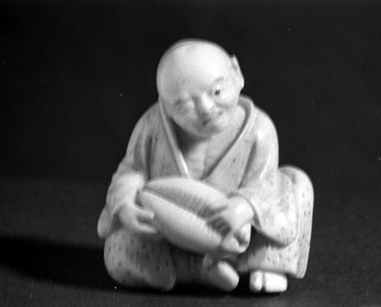 Hearst Museum object titled Netsuke, accession number 9-7536, described as Netsuke: old man seated and holding cowrie shell. His robe elaborately designed and decorated. ••According to the donor's catalog: "Netsuke in ivory of a blind old man, seated and holding a cowrie shell. They are a very beautiful shell and are said to have been used as coinage as far back as the Chou Dynasty and continued in circulation as late as the fourteenth century. Wu Ti of 140 until 87 BC of the Han Dynasty was very active in his regulation of the coinage system and the Chou long preceded him—circa 1136 to 250 BC. The old blind man of our netsuke is in possession of a very superior cowrie and his sensitive fingers of a blind man have been able to recognize the beauty and coinage value of the shell, and he is saying to himself with an accentuating wink, 'I have the Kale!'