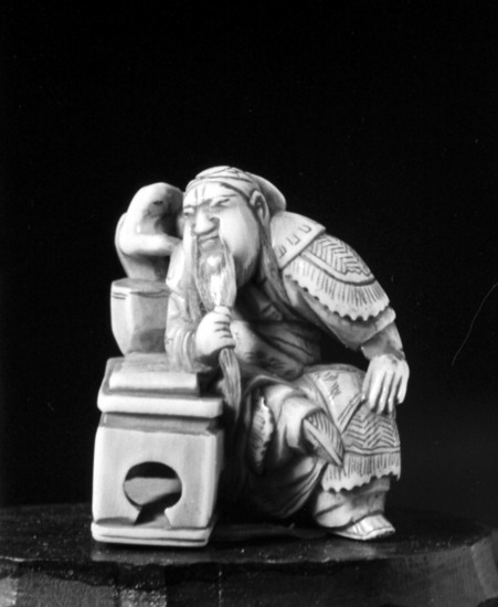 Hearst Museum object titled Netsuke, accession number 9-7529, described as Netsuke: man with long beard seated at table. Scroll and vase containing coral branch on table.