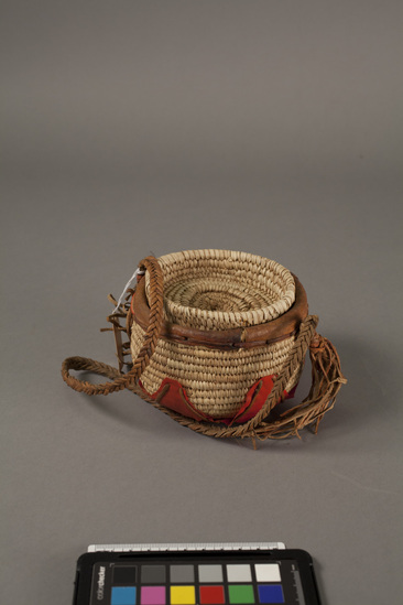 Hearst Museum object titled Basket with lid, accession number 5-10459a,b, described as Container; palm fiber with leather rim and red plastic covering base. Lid; palm fiber, diameter 12 cm. Leather plaited cord, ca length 56 cm, height 11 cm. A: container. B: Lid. Native name: Omra.