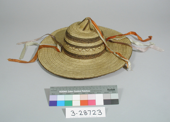 Hearst Museum object titled Woven straw hat with ribbon, accession number 3-28723, described as Hat. Wide flat brim with small cone shaped piece attached to top of crown. Plaited strips of palm leaf sewn together by hand, some dark strips in crown and one around outer under edge; white cotton string chin strap. Faded red, white, and blue ribbons attached at sides of crown and on top of cone piece. Diameter: 39 cm.