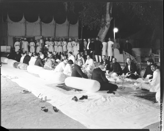 Hearst Museum object titled Black-and-white negative, accession number 15-30483, described as 8 x 10" Black and white negative of a banquet with The Nawab of Bhopal at Camp Dewatya. Hoefler's original description: "Bhopal State, Central India. A banquet in the Indian style, given by His Highness The Nawab of Bhopal at Camo Dewatya. A properly prepared Indian dinner, such as this one, is a delight to the epicure. His Highness of Bhopal is noted, not only for his hospitality, but also for the excellency of his table, this being due, in a large measure, to the fact that he has an English Butler who supervises every detail of his household, including his kitchen. No wines or liquors of any kind are served at the table of His Highness, for he is a Mohammedan and a strict teetotaler. There are many notable people at the table, but suffice to say that the Gentleman in the center, between the two ladies, is His Highness The Nawab.