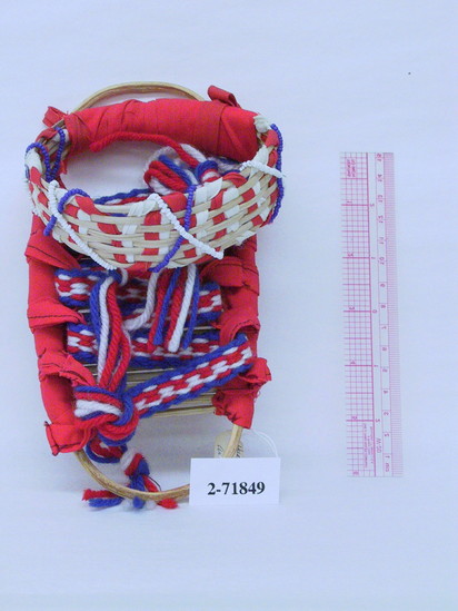 Hearst Museum object titled Toy cradle board, accession number 2-71849, described as Hualapai toy cradle board.  Broad is of willow sticks attached to an oval frame with a full hoop head shade, decorated with red and white cotton strips and blue and white strings of beads.  Sides and shade have red cotton winding and straps are red, white & blue braided yarn.    L = 7.75”