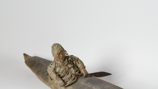 Hearst Museum object titled Kayak model, accession number 2-4568, described as 2-hatch skin boat (ulluxtadaq) model with one wood human figure with gut clothing. Gut cover, wood frame. Remnants of red and blue yarn on central deck seam. One miniature paddle.