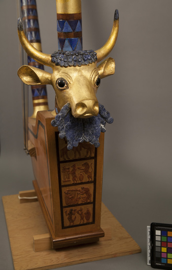 Hearst Museum object 2 of 3 titled Lyre, accession number 9-12847, described as Harp reproduction; soundbox Bird's Eye maple; two mahogany posts inlaid with 288 pieces of blue enamel on copper; on front post is wood bull's head overlaid with 23K gold leaf; bull's beard, forehead and horn-tips blue/white sodalite, obsidian eyes; below bull's head are four low relief panels of contemporary scenes; on rear post are 2 upright goats in low relief; crossbar inlaid with single blue/white sodalite at each end; 13 pegs above, 12 strings; height 1.19 m to top of pegs, length 91.5 cm (crossbar).  Reproduction of Sumerian specimen (made in Quincy, Mass. by Eli Richman). Original harp excavated by Sir Leonard Wooley from Ur, Mesopotamia, circa 3,000 BC. Original is no. U.10556 and is in the University Museum, Philadelphia (Kilmer).