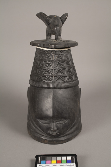 Hearst Museum object 4 of 4 titled Helmet mask, accession number 5-6260, described as Helmet mask of black stained gum wood; at bast in front is carved face; top half if five horizontal rows of incised triangles; at top is bird carved in the round. Height 43 cm.