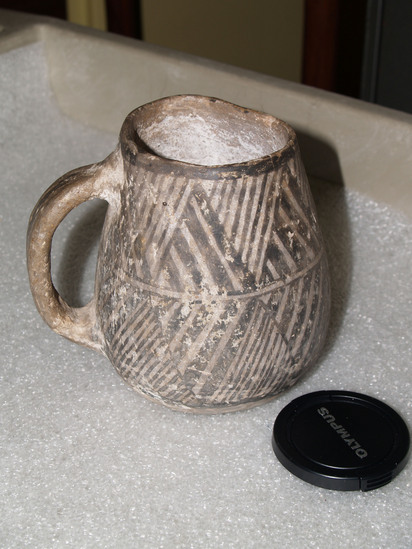 Hearst Museum object 1 of 3 titled Mug, accession number 2-65925, described as Mug with single handle; buff pottery; 2 bands of zigzags with diagonal fillers around the exterior.  The rim is chipped.  Pueblo III per Larry Dawson.