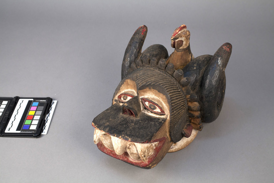 Hearst Museum object titled Head mask, accession number 5-3820, described as Carved wooden head mask; Olágá; dog-face with curved ram horns; chicken effigy on top; red, white, and black; 12 3/2 inches x 8¼ inches.