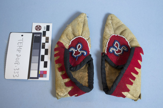 Hearst Museum object titled Mocassins, accession number EOC2019.380, described as Pair of child's moccasins, made of leather, red and black felt, and decorated with small white, blue, and red beads. Slightly squashed and misshapen.