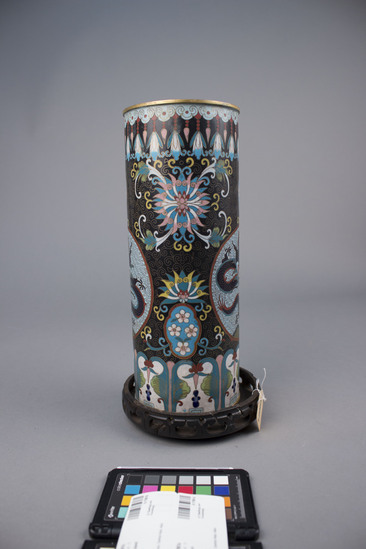 Hearst Museum object titled Urn, enamel on metal, multicolored, accession number 9-17861a,b, described as (a) Vase, tall cylindrical, enamel on brass.  Exterior cloisonne: two central medallion depicting a dark blue, wire scaled, “5-toed” dragon with turquoise and red accents on light blue ground with stylized wire cloud diaper; stylized, multicolored lotuses and small light blue scallop-shaped medallions with white quadrefoil flowers, all on a black ground with wire; wire scrolls surrounds medallions; at base and rim, band of multicolored stylized flowers, rim black ground, base white ground. (b) circular teakwood stand with a single band of carved thunder clouds around side