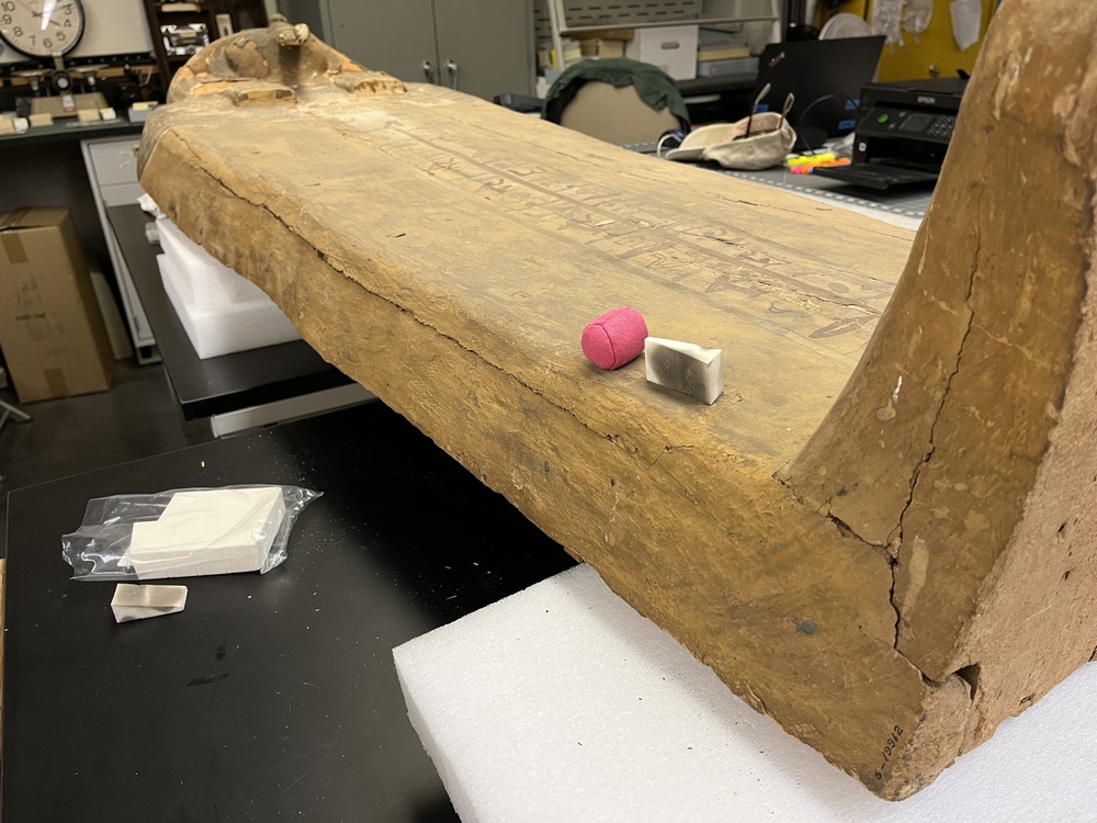 Hearst Museum object 19 of 167 titled Coffin, accession number 6-19912, described as Wooden anthropoid coffin. Inscription running down center of lid. Determined by Kea Johnston to probably be the outer coffin of Iwefaa (6-19928, 12-11039). According to Kea Johnston, "This coffin belonged to a man named Iwefaa, whose name is inserted in the middle of the spells written in the two columns on the front. This coffin is likely the outer coffin for PAHMA 6-19928 based on the similarity of the owner's name, unusual costume, and the paleography of the inscription. Both coffins were purchased on the antiquities market by George Reisner for Phoebe A Hearst. However, stylistically, they are very similar to a set of coffins known to have come from the site of Akhmim, which was excavated in the 1880s by Gaston Maspero, and which has heavily looted for the antiquities market during this decade." "The pectoral vignette between the text and the collar shows the deceased standing before squatting baboon and is the source of our logo. The hieroglyph for 'sky' is above their heads, and the side borders are formed by a pyramid and the symbol for the West.