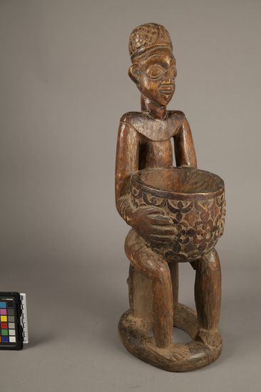 Hearst Museum object 3 of 4 titled Carved wood figure, accession number 5-6001, described as Wood male figure seated on stool; holding bowl with human-like figures in low relief; stool is carved human head; h. 63.5 cm. Fine brown patina; some termite damage on base.