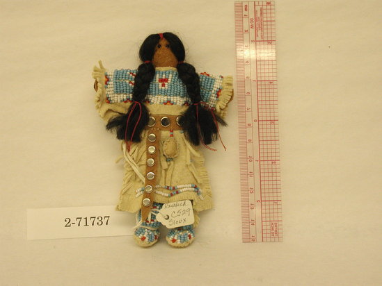 Hearst Museum object titled Doll, accession number 2-71737, described as Female doll. Buckskin body with beaded eyes, black embroidery floss hair in braids tied with red thread.  Two-skin dress heavily beaded in blue and white with red accents.  Turtle amulet contains sage. Leather belt with conchos.  Beaded moccasins.  H = 5.5”