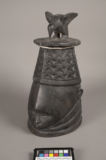 Hearst Museum object titled Helmet mask, accession number 5-6260, described as Helmet mask of black stained gum wood; at bast in front is carved face; top half if five horizontal rows of incised triangles; at top is bird carved in the round. Height 43 cm.