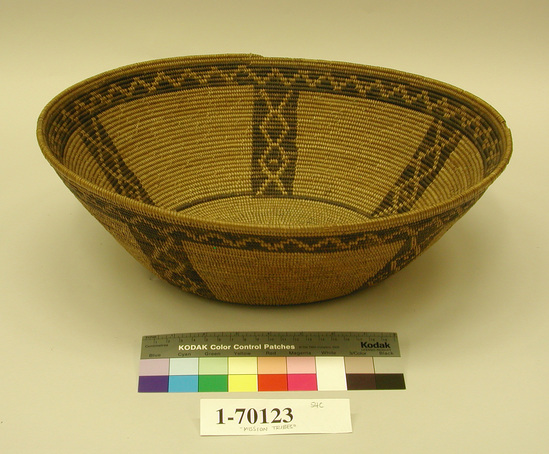 Hearst Museum object titled Cooking basket, accession number 1-70123, described as Coiled, deep tray.  Tag "Mission Type". Per Ralph Shanks:  Broad bowl in cooking basket shape, possibly used.  The basket has an oval start which is unusual in a round basket like this.  Wear patterns on the basket make it appear old.  The materials include a deergrass bundle foundation; sumac weft on the base and sides with tan and black-dyed juncus designs.  The design is a primary band of zigzags.  The design is two coils below the rim.  There are five vertical bands of chains of diamonds on the sides of the basket and one coil of black-dyed juncus on the beginning of the base.   Workface is on the interior.  Rightward warp direction with clipped and bound under weft fag ends.  The clipped fag ends are used along the edges of the designs and in the designs.  There are some clipped fag ends in the background.  The bound under fag ends are primarily in the background.  The basket has a plain-wrapped rim with an inch and a half herringbone ending.   Attributed to the Gabrielino because of the design layout, which includes the primary design two coils below the rim, the vertical designs ending at the base with a coil of black-dyed juncus, the presence of clipped fag ends, and the use of a sumac in the base.  The basket design layout is only used by the Gabrielino and the Chumash.  The Chumash place their design further away from the rim and have less use of bound under weft fag ends, and are more likely to use a three rod juncus foundation.