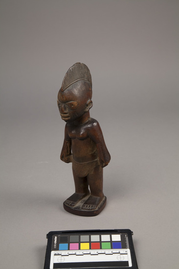 Hearst Museum object 6 of 6 titled Ere ibeji figure, accession number 5-15807, described as Figure, twin (Ibeji), wood, handcarved; female, with elongated headdress, wearing grass skirt; hands on hips; straight legs, with feet mounted on keystone-shaped base; dark brown, polished finish. Bottom stamped "Product of the Gold Coast" by William Bascom in compliance with customs regulation, per collector's notes.